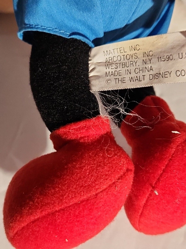 K&ouml;nnte beinhalten: Ein schwarzes und rotes Pl&uuml;schtier mit einem blauen Hemd. Das Spielzeug hat ein Etikett mit der Aufschrift "MATTEL INC. ARCO TOYS, INC. WESTBURY, N.Y. 11590, U.S. MADE IN CHINA &copy; THE WALT DISNEY CO."