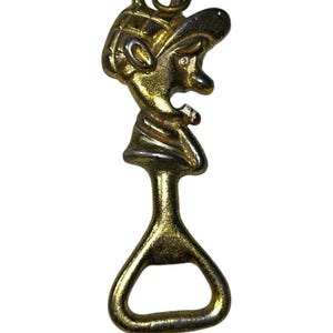 May include: A vintage bottle opener with a gold-toned finish. The opener features a stylized figure with a hat and open mouth, and a small red accent. The handle has a teardrop shape.