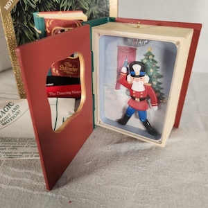 May include: Hallmark Keepsake ornament featuring a miniature scene from The Nutcracker ballet. The ornament is shaped like a book, with a red cover and a diorama inside depicting a toy soldier, Christmas tree, and disco ball. The box reads "Magic Light! Motion! Music!"