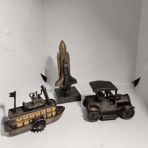Könnte beinhalten: Drei vintage Bleistiftspitzer aus Metall: ein Raumschiff, ein Auto und ein Schaufelraddampfer. Das Raumschiff ist golden, das Auto ist schwarz und das Boot ist braun mit gelben Akzenten.