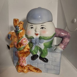 May include: A ceramic teapot shaped like a person wearing a blue hat and sitting on a brick wall. The teapot has a green and purple outfit and is holding a flower-covered branch.