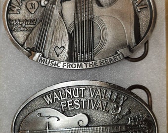 May include: Two silver belt buckles with a musical theme. The top buckle features a guitar and the text "WALNUT VALLEY FESTIVAL 1972-2002 MUSIC FROM THE HEART". The bottom buckle features a mandolin and the text "WALNUT VALLEY FESTIVAL WINFIELD, KANSAS 2003".