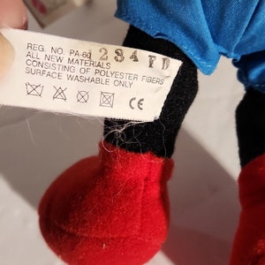 K&ouml;nnte beinhalten: Ein schwarzes und rotes Pl&uuml;schtier mit einem blauen Hemd. Das Spielzeug hat ein Etikett mit der Aufschrift "REG. NO. PA-60 284 FD ALL NEW MATERIALS CONSISTING OF POLYESTER FIBERS SURFACE WASHABLE ONLY XXCE".
