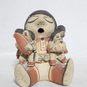 Pode incluir: Estatueta de cerâmica que retrata uma figura sentada com a boca aberta, rodeada por figuras menores. A estatueta é pintada em tons de creme, castanho e laranja, com detalhes nas roupas e rostos. O design sugere um estilo de arte popular.