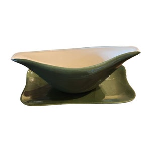 May include: A ceramic gravy boat and matching saucer. The boat is a deep green with a white interior and a curved, boat-like shape. The saucer is a matching green with a curved design.