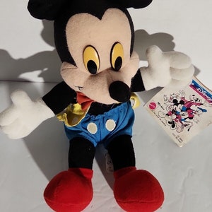 K&ouml;nnte beinhalten: Ein Pl&uuml;sch-Mickey-Mouse-Spielzeug, das ein gelbes und blaues Outfit mit roten Schuhen tr&auml;gt. Das Spielzeug sitzt auf einer wei&szlig;en Oberfl&auml;che mit einem Etikett, auf dem "Disney's Mickey Mouse" steht.