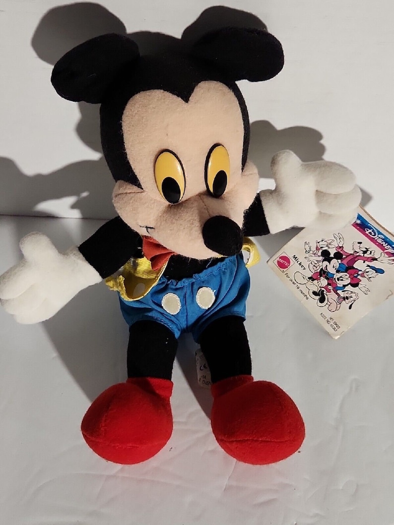 K&ouml;nnte beinhalten: Ein Pl&uuml;sch-Mickey-Mouse-Spielzeug, das ein gelbes und blaues Outfit mit roten Schuhen tr&auml;gt. Das Spielzeug sitzt auf einer wei&szlig;en Oberfl&auml;che mit einem Etikett, auf dem "Disney's Mickey Mouse" steht.