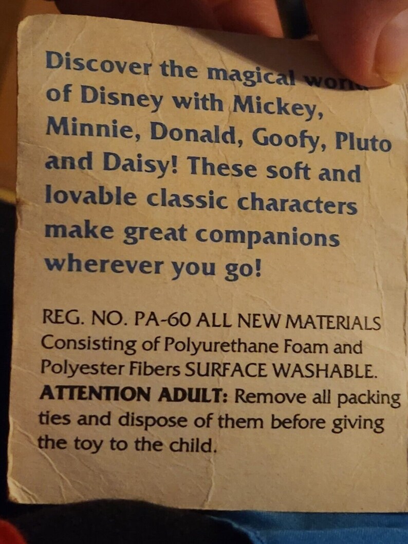 K&ouml;nnte beinhalten: Nahaufnahme eines Produktetiketts mit dem Text "Entdecken Sie die magische Welt von Disney mit Mickey, Minnie, Donald, Goofy, Pluto und Daisy! Diese weichen und liebenswerten klassischen Figuren sind gro&szlig;artige Begleiter, wohin Sie auch gehen! REG. NO. PA-60 ALL NEW MATERIALS Consisting of Polyurethane Foam and Polyester Fibres SURFACE WASHABLE. ATTENTION ADULT: Remove all packing ties and dispose of them before giving the toy to the child."