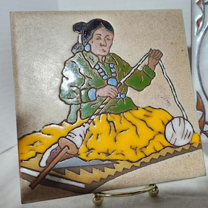 May include: Square ceramic tile featuring a Native American woman in a green jacket and yellow skirt, seated and working with yarn. The artwork is outlined in black, with a cream-colored background and a decorative border. The tile is displayed on a gold-colored stand.