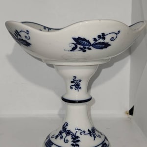 Puede incluir: Un cuenco de cerámica blanca con un diseño floral azul sobre un pedestal. El cuenco tiene un borde festoneado y se asienta sobre un pedestal alto y delgado.