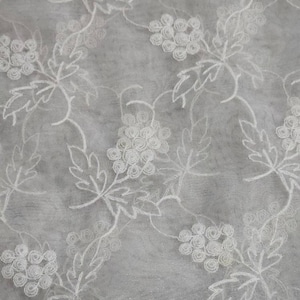 May include: Close-up of a sheer, white fabric with an embroidered floral pattern. The design features clusters of flowers and leaves, creating a delicate and textured surface. The background is a soft, light gray.