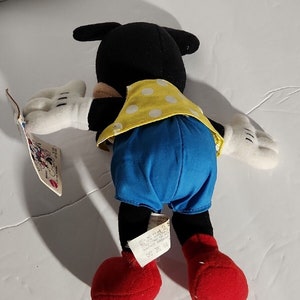 K&ouml;nnte beinhalten: Ein schwarzes und wei&szlig;es Pl&uuml;sch-Mickey-Mouse-Spielzeug, das ein gelbes und wei&szlig;es gepunktetes Hemd und blaue Shorts tr&auml;gt. Das Spielzeug liegt auf dem R&uuml;cken mit ausgestreckten Armen und gebeugten Beinen. Das Spielzeug hat ein rotes Etikett an seinem Fu&szlig; mit dem Text "Mickey Mouse" und "Hergestellt in China".