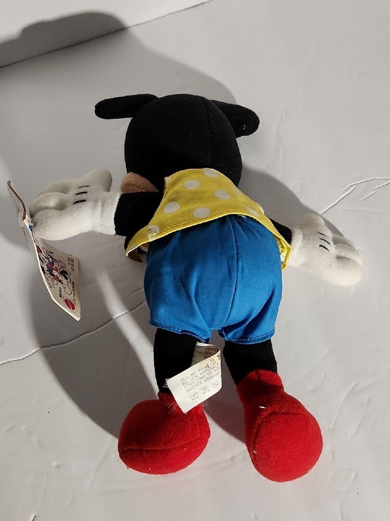 K&ouml;nnte beinhalten: Ein schwarzes und wei&szlig;es Pl&uuml;sch-Mickey-Mouse-Spielzeug, das ein gelbes und wei&szlig;es gepunktetes Hemd und blaue Shorts tr&auml;gt. Das Spielzeug liegt auf dem R&uuml;cken mit ausgestreckten Armen und gebeugten Beinen. Das Spielzeug hat ein rotes Etikett an seinem Fu&szlig; mit dem Text "Mickey Mouse" und "Hergestellt in China".