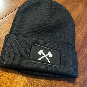 以下が含まれることがあります： 白の交差した斧のデザインが特徴の長方形のパッチが付いた黒いビーニー帽。帽子はニット素材でできており、折り返しのつばが付いています。帽子は無地の黒色です。