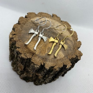 May include: Two pairs of axe earrings, one pair is silver and the other is gold. The earrings are displayed on a wooden surface.