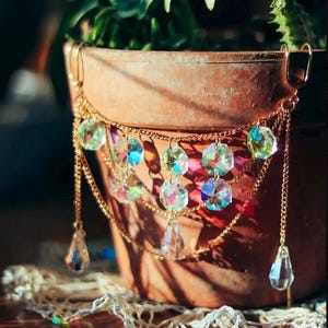 May include: A gold-colored metal plant pot hanger with iridescent crystal accents. The hanger features multiple strands of delicate chains and teardrop-shaped crystals. The crystals reflect a rainbow of colors, adding a touch of sparkle to the terracotta pot.
