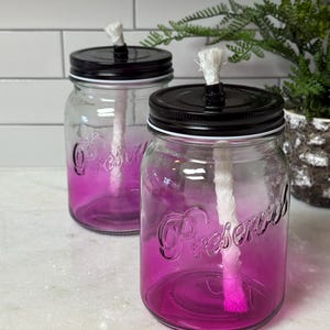 Pode incluir: Duas lâmpadas de óleo de vidro em estilo jarro Mason com tampas pretas e pavios brancos. Os frascos têm uma cor rosa gradiente e a palavra "Preserved" está em relevo na frente. As lâmpadas estão em uma superfície branca.