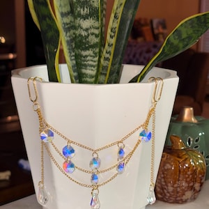 May include: A gold-colored chain plant hanger with iridescent crystal accents. The plant hanger is suspended from a white square pot containing a green plant. The crystals reflect multiple colors.