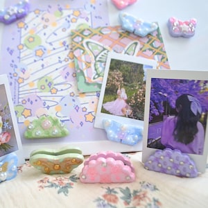 May include: A collection of decorative photo holders in various pastel colors, including blue, pink, green, and purple. Each holder is shaped like a butterfly or a scalloped design and embellished with pearls and rhinestones. The holders are supporting polaroid-style photos.