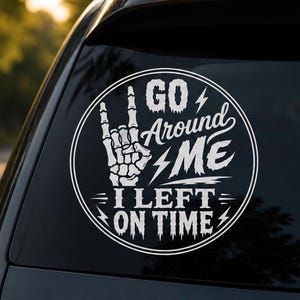 Può includere: Un adesivo circolare in vinile bianco applicato sul lunotto posteriore di un'auto. Il design presenta una mano di scheletro che fa un gesto rock-and-roll accanto al testo GO AROUND ME I LEFT ON TIME, con fulmini e un bordo circolare.