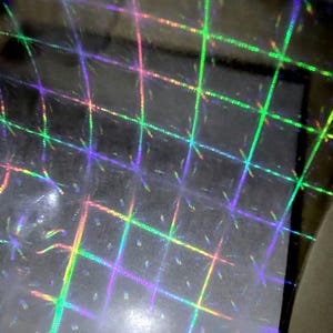 May include: Close-up of a holographic sheet with a grid pattern. The grid lines are iridescent, displaying a spectrum of colours including green, purple, and orange. The background is a dark, reflective surface.