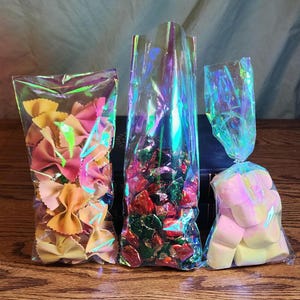 May include: Three iridescent cellophane bags filled with different treats. The bag on the left contains colorful bowtie pasta, the middle bag contains red and green wrapped candies, and the bag on the right contains white and yellow marshmallows.