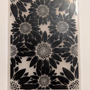 Può includere: Una cartella per goffratura rettangolare trasparente con un motivo ripetuto di girasoli neri. I girasoli hanno petali strutturati e capolini centrali, creando un effetto dimensionale. Il design è su uno sfondo traslucido.