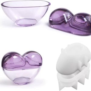Puede incluir: Cuatro objetos de vidrio: un cuenco ovalado transparente, una forma de doble burbuja de vidrio púrpura, un recipiente de vidrio en forma de corazón con tapa púrpura y un molde blanco con una forma única. Las piezas de vidrio tienen un acabado liso y brillante.