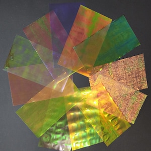 Puede incluir: Surtido de láminas holográficas iridiscentes en varios colores, incluyendo verde, dorado, naranja y morado. Las láminas están dispuestas en un patrón circular.