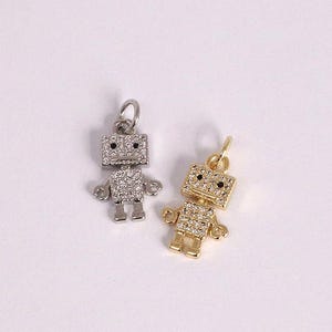 May include: Two robot-shaped charms, one silver-toned and one gold-toned, are displayed against a white background. Each charm is encrusted with small, clear crystals, with black accents for the eyes. The charms are designed for jewelry making.