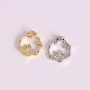 Può includere: Due ciondoli, uno dorato e uno argentato, ciascuno con una cornice trasparente dai bordi ondulati. All'interno, è visibile un motivo a forma di sole con piccole stelle. I ciondoli sono probabilmente per la creazione di gioielli o l'artigianato.