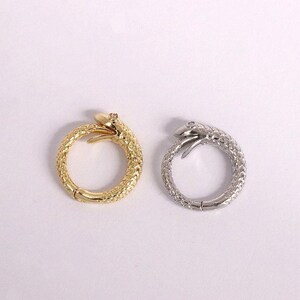 Pode incluir: Dois anéis em forma de cobra, um dourado e outro prateado, são exibidos sobre um fundo branco. Os anéis apresentam um padrão texturizado em forma de escamas e um design de cabeça de cobra. O anel dourado tem uma pequena gema na cabeça da cobra.