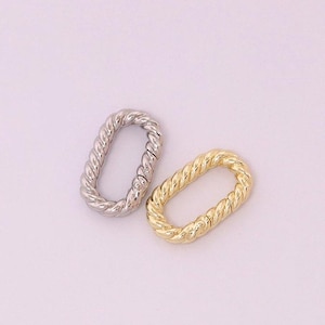 May include: Two oval-shaped jewellery clasps. One is silver-coloured, and the other is gold-coloured. Both clasps have a twisted rope design. The clasps are displayed on a light pink background.