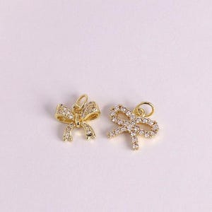 Könnte beinhalten: Zwei goldfarbene Schleifenanhänger, jeweils mit kleinen, klaren Kristallen verziert. Die Schleifen haben ein zartes, feminines Design mit einer Schlaufe oben zum Befestigen an Schmuck. Die Anhänger sind vor einem weichen, cremefarbenen Hintergrund platziert.