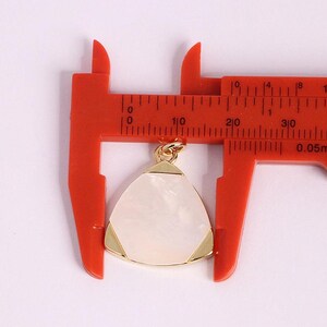 May include: A gold-toned pendant with a pearlescent, triangular center is displayed against a red caliper. The pendant's corners are accented with gold. The caliper shows measurements in millimeters.