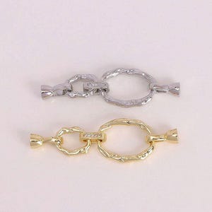 May include: Two decorative chain links, one silver and one gold, are displayed against a white background. Each link features an oval-shaped center with a textured surface and a smaller, connecting ring with a row of small, clear stones.