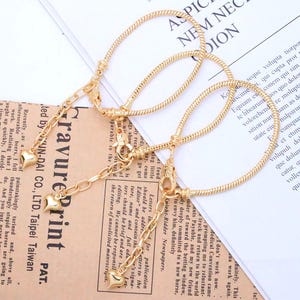 Puede incluir: Pulseras doradas con diseño de cadena de serpiente. Cada pulsera presenta una cadena con dijes en forma de corazón y un cierre de mosquetón. Las pulseras se exhiben sobre un fondo de periódico.