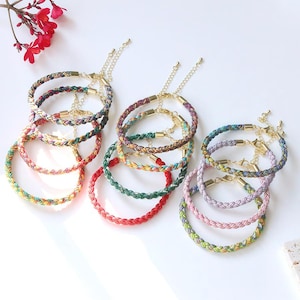 Könnte beinhalten: Eine Sammlung bunter geflochtener Armbänder mit goldfarbenen Verschlüssen. Die Armbänder weisen eine Vielzahl von Farben auf, darunter Rot, Grün, Lila und Gelb. Die Armbänder werden auf einer weißen Oberfläche präsentiert.