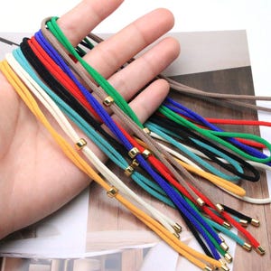 May include: A collection of multi-coloured cord necklaces in yellow, white, turquoise, black, red, blue, green, and beige. Each cord features a small, polished gold-tone metal bead accent that slides along the length of the fabric material.