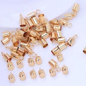 Peut inclure: Une collection de petits embouts à sertir de couleur or brillant avec des boucles attachées. Ces apprêts de bijoux sont cylindriques et en forme de cloche, conçus pour fixer les extrémités des cordons ou des chaînes en bijouterie. Les embouts sont dispersés sur un fond clair.