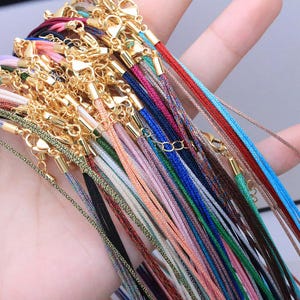Peut inclure: Une collection de colliers en cordon colorés avec des fermoirs dorés. Les cordons sont de différentes couleurs, notamment rouge, bleu, vert, rose et noir. Les colliers sont présentés ensemble, mettant en valeur la variété des couleurs et des designs.