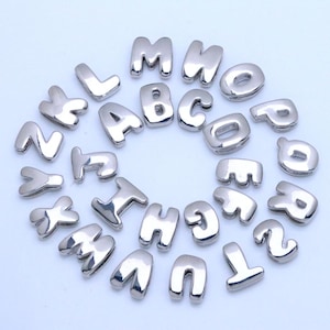 Op de afbeelding: Zilverkleurige alfabetletterbedels in een afgeronde stijl. De letters zijn in een verspreid patroon gerangschikt. De letters zijn gemaakt van een glanzend, metallic materiaal. Geschikt voor het maken van sieraden.