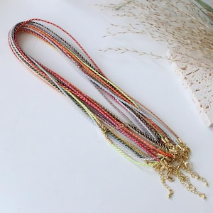 Puede incluir: Una colección de collares trenzados multicolores con cierres y cadenas de extensión doradas. Los collares presentan una variedad de colores, incluyendo rojo, naranja, amarillo, verde y plateado. Los collares se muestran sobre una superficie blanca.