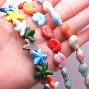 May include: Two strands of ceramic beads shaped like marine life. One strand features colourful starfish in yellow, white, green, grey, and blue. The other strand consists of various spiral seashells in shades of orange, white, red, pink, and light green.