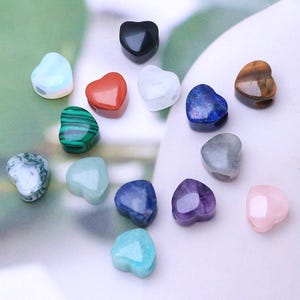 Puede incluir: Una colección de piedras preciosas en forma de corazón en varios colores y texturas. El surtido incluye piedras en tonos de azul, verde, rojo, morado y marrón. Las piedras preciosas están dispuestas sobre una superficie clara, mostrando sus patrones y tonalidades únicos.