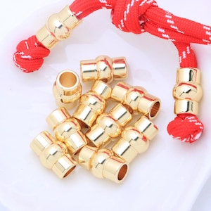 May include: Gold-coloured metal beads and cord ends. The beads are cylindrical with a rounded centre and are scattered on a white surface. Red and white braided cord is attached to the gold beads.