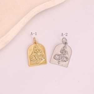 Peut inclure: Deux breloques de pendentif en métal. L'une est dorée, l'autre argentée. Chaque breloque présente un motif de serpent dans une forme d'arche rectangulaire. Les breloques sont étiquetées A-1 et A-2.