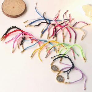 May include: A collection of colorful braided bracelets with gold-colored beads and accents. The bracelets feature various colors, including red, pink, blue, yellow, and green. Some bracelets have a circular pendant with a design.