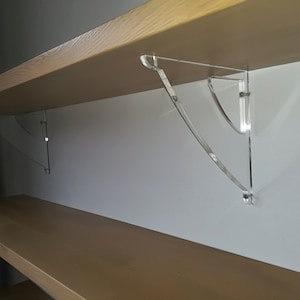 Pode incluir: Dois suportes de prateleira de acrílico transparente são montados em uma parede branca, sustentando uma prateleira de madeira. Os suportes são curvos e têm um design moderno.