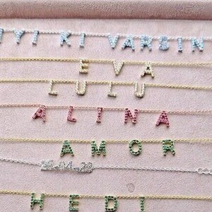 Peut inclure: Un présentoir de colliers avec différents noms écrits en chaînes dorées et argentées avec des pierres précieuses colorées. Les noms incluent Iyi, Ki, Varsin, Eva, Lulu, Alina, Amor, Hedi, Fatumata, Briana et Aju.